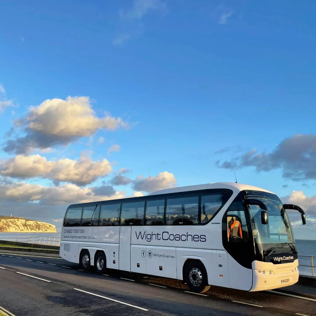 White Wight Coaches bus on a coastal road under a partly cloudy blue sky.