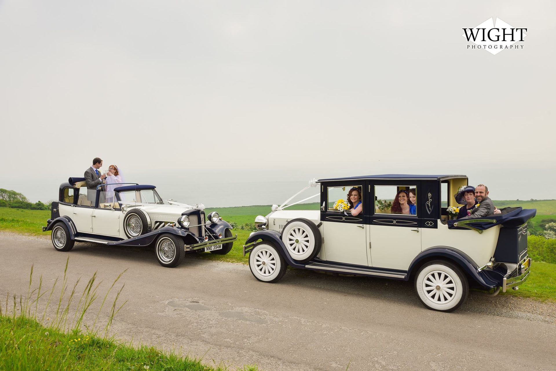 Two vintage white cars with wedding party on a road overlooking a landscape.