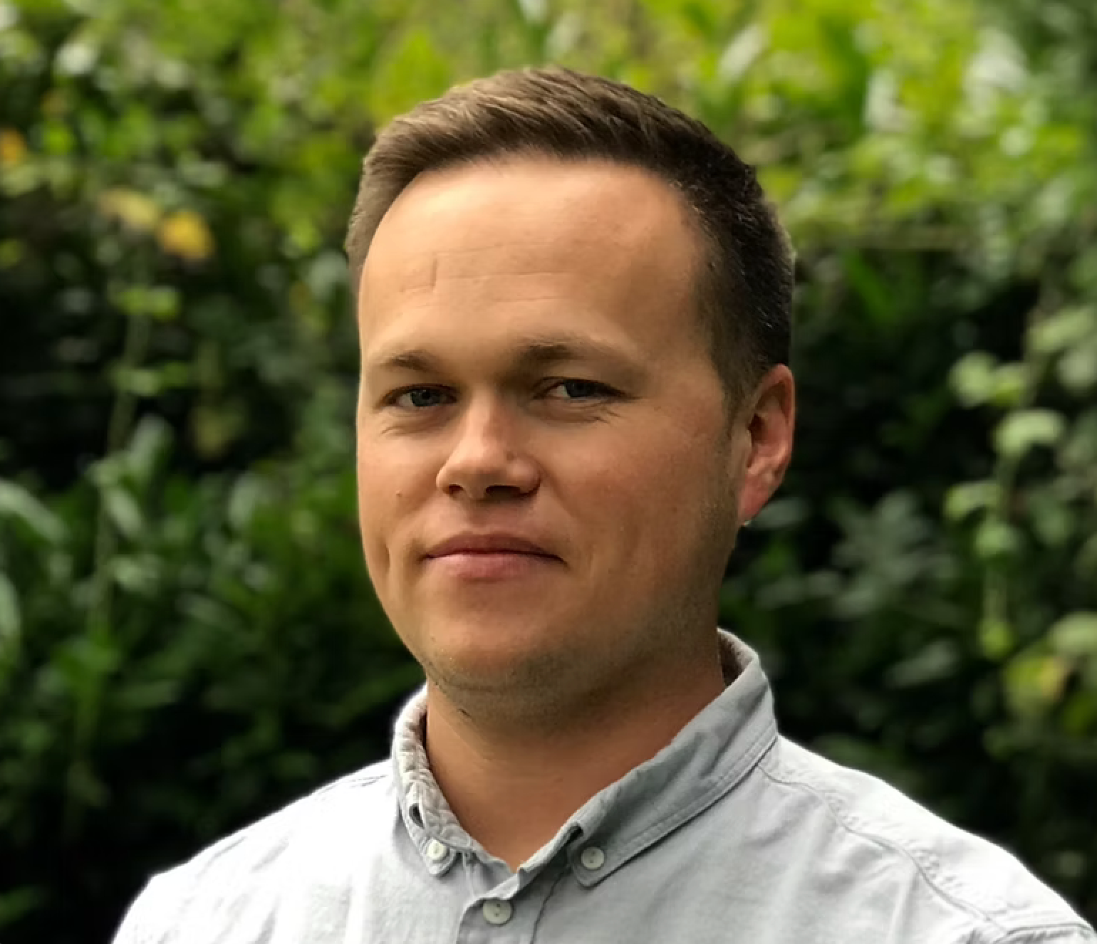 A man in a white shirt is standing in front of a bush.