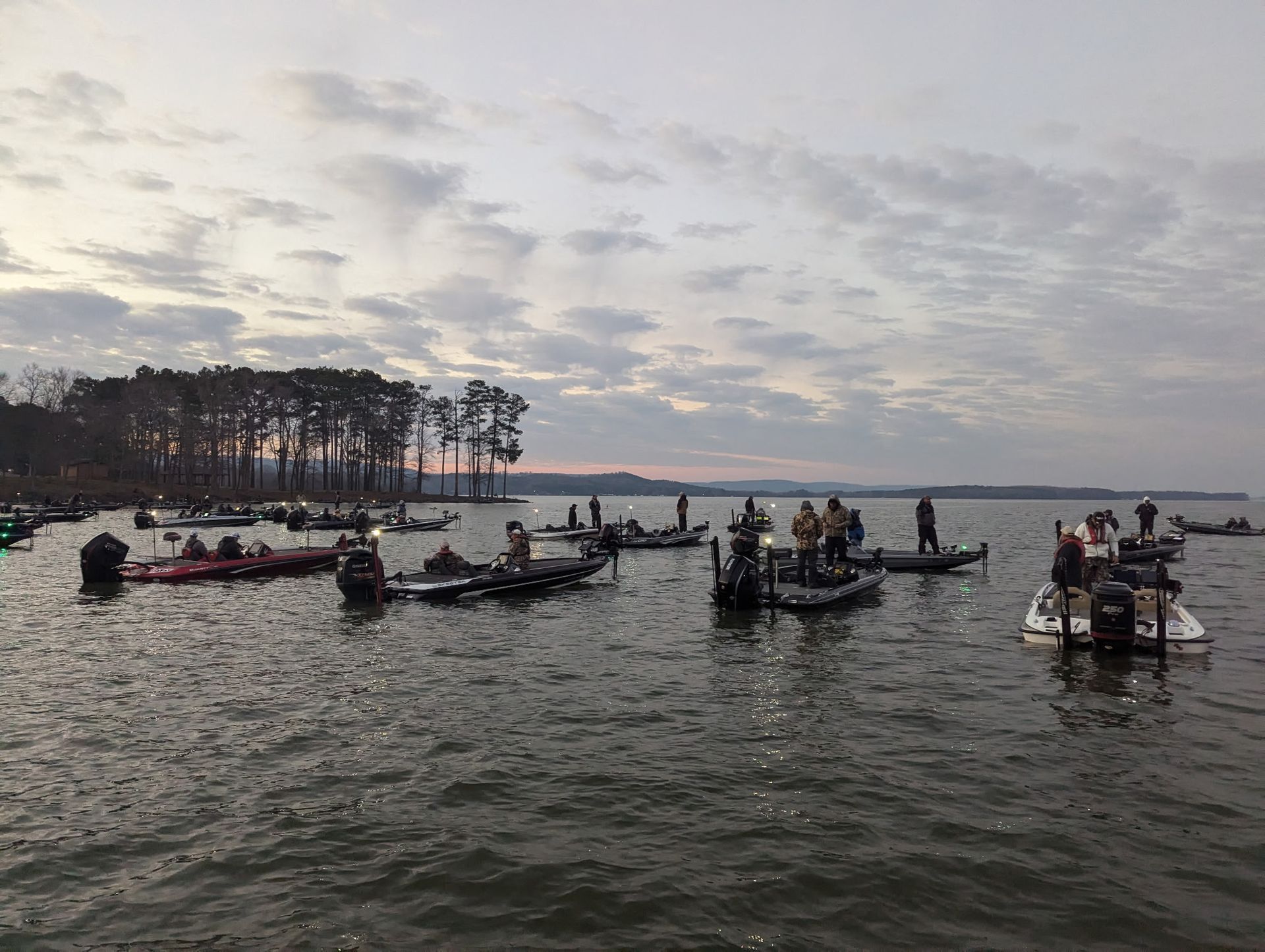 Bass boats on a calm lake at dusk, with a tree-lined shore and cloudy sky.