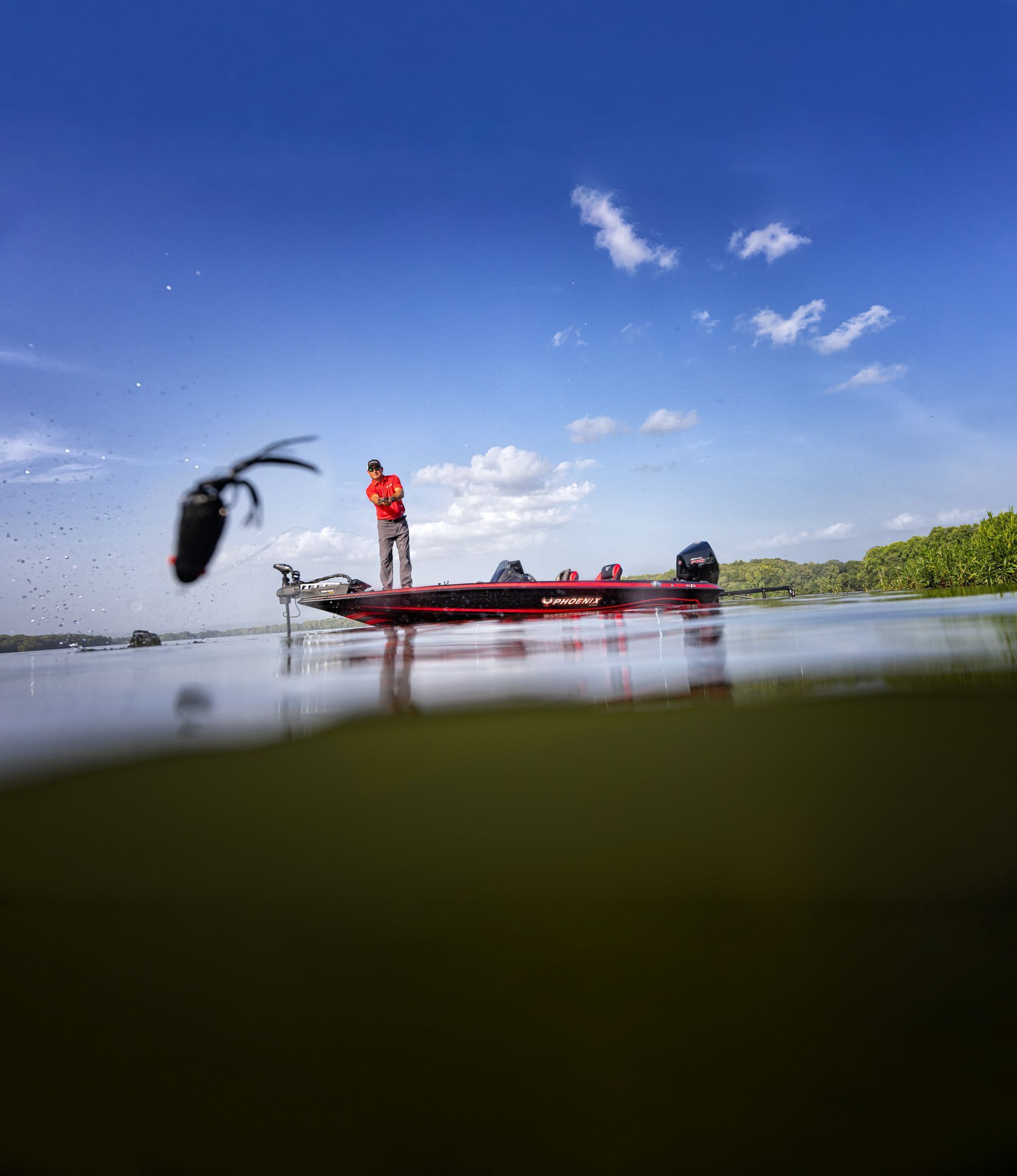 Angler on a red boat under a blue sky, fishing line cast over calm water near green shoreline.