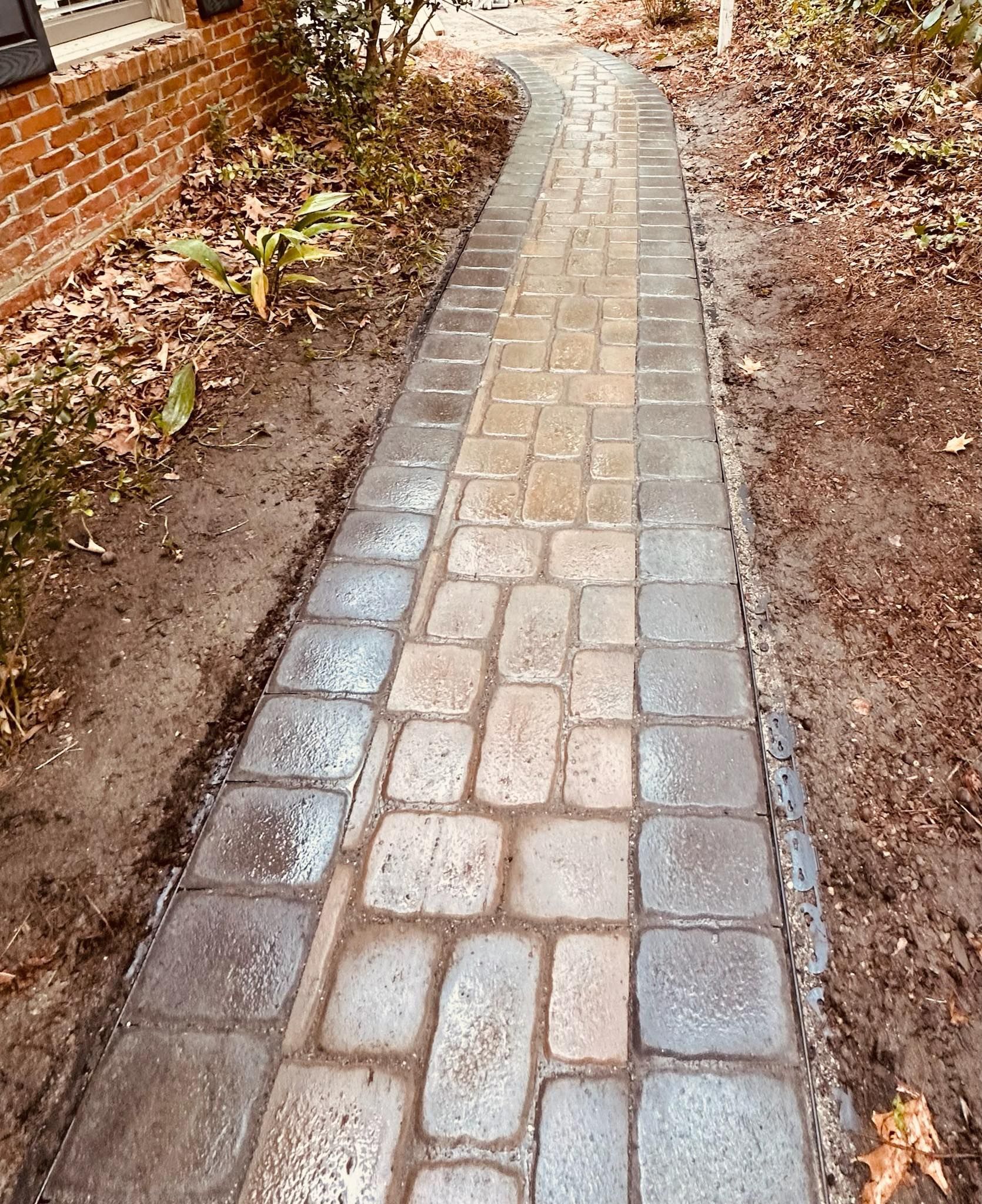 A narrow stone walkway bordered by dirt and fallen leaves beside a brick wall.