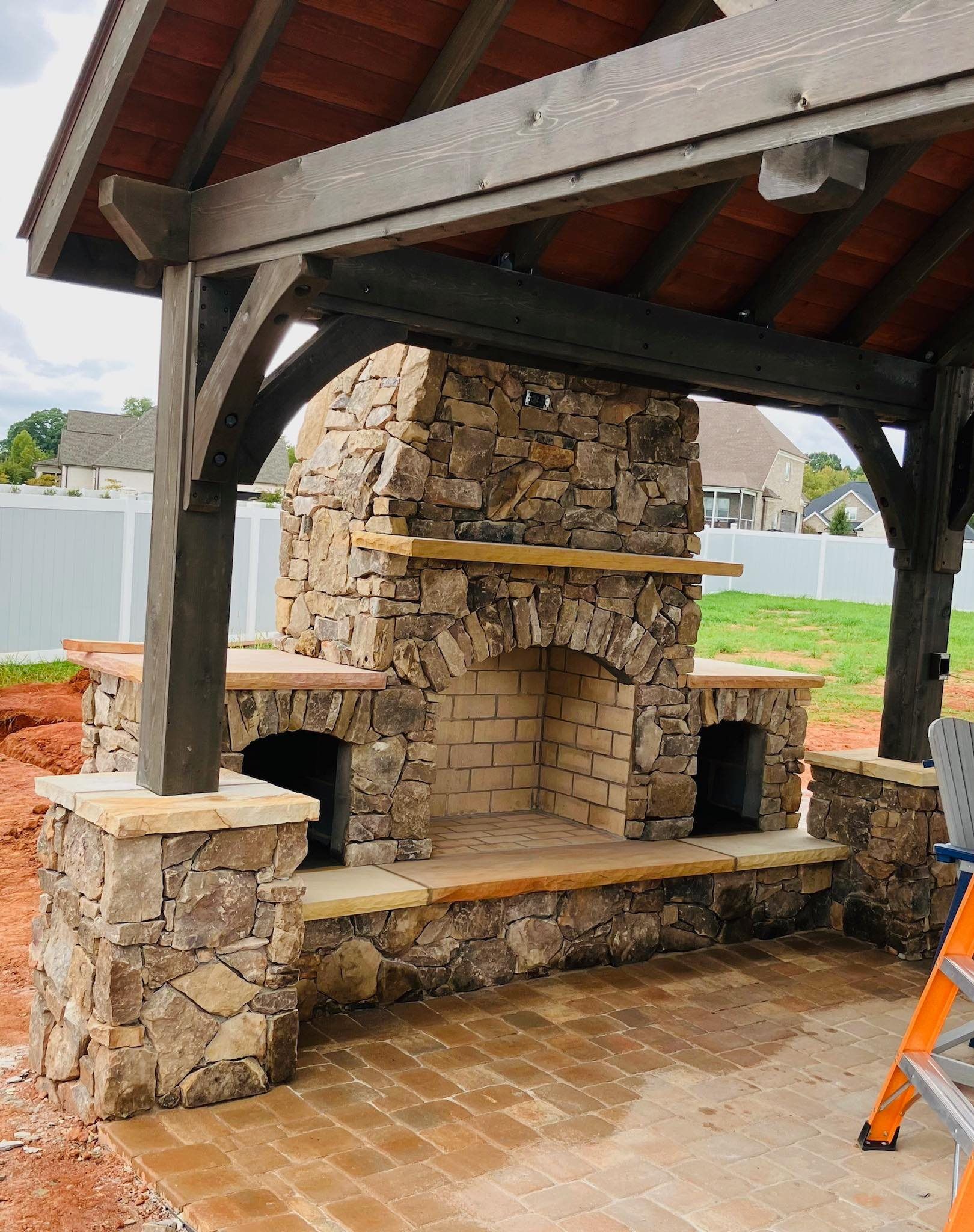 Stone outdoor fireplace under a covered patio with a wood roof and brick floor