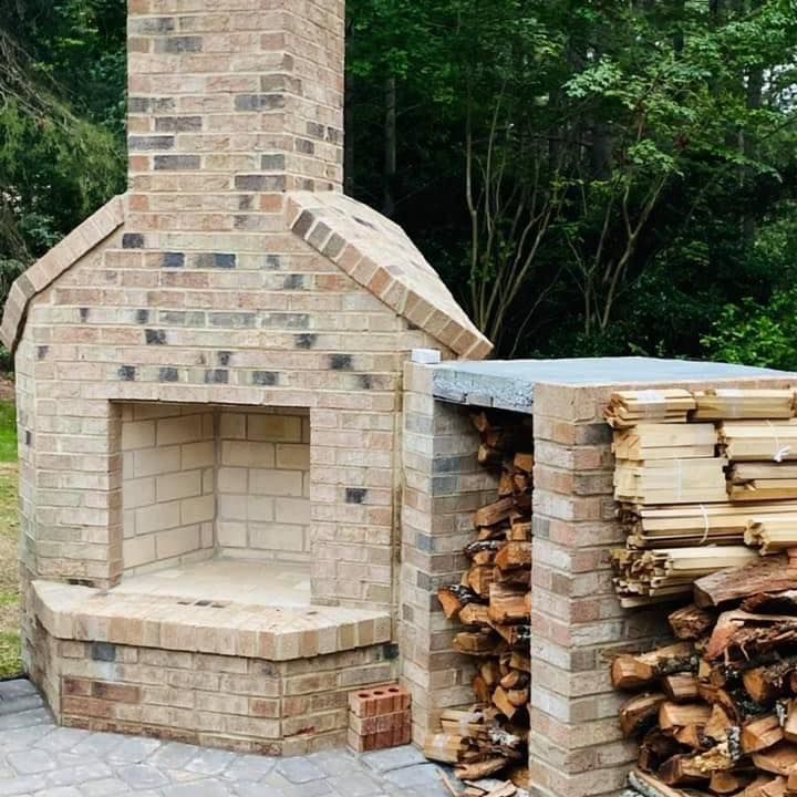 Brick outdoor fireplace beside stacked firewood in a wooded yard
