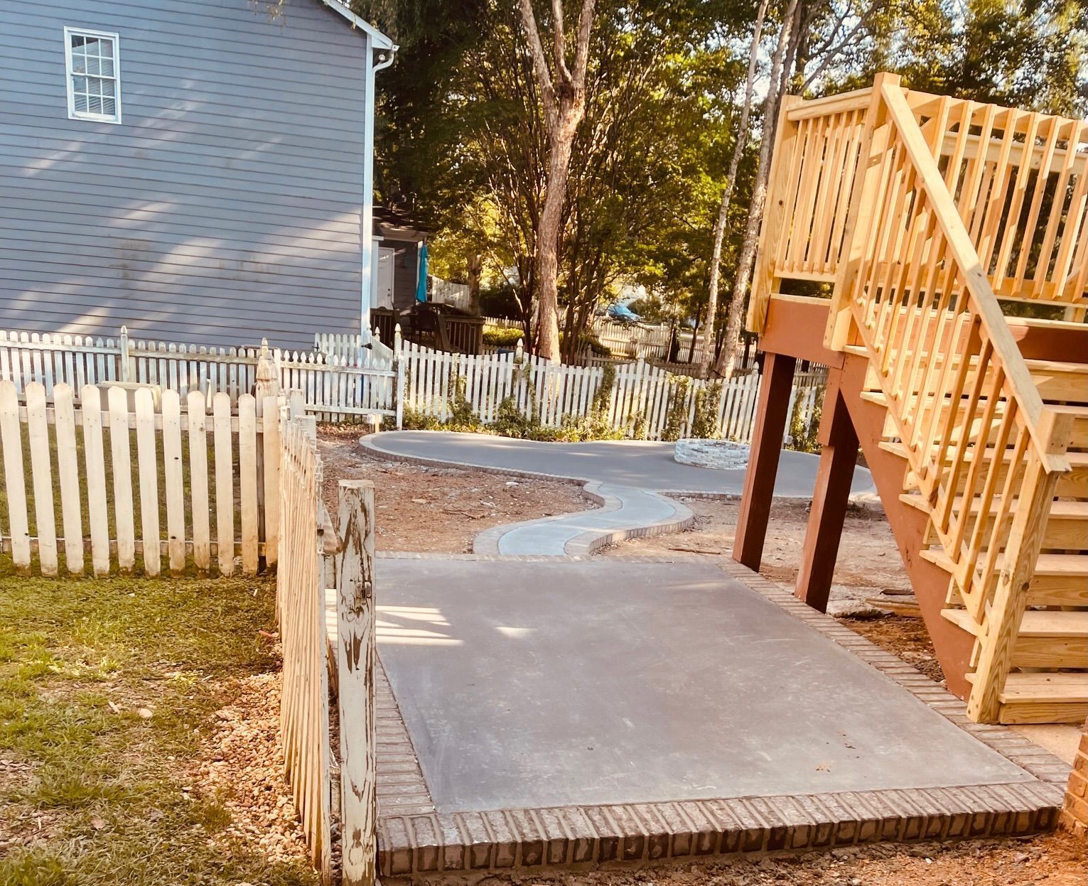 Backyard patio with concrete walkway, white fence, and wooden stairs beside a house