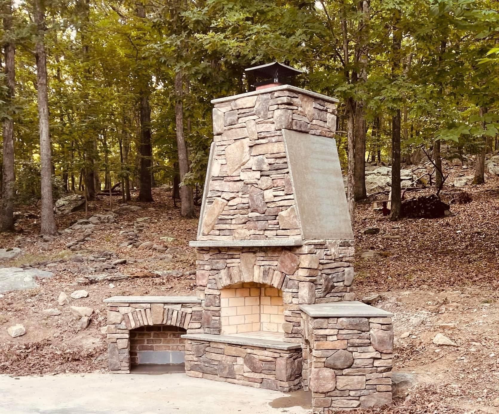 Stone outdoor fireplace with chimney in a wooded area, set against fallen leaves and trees.