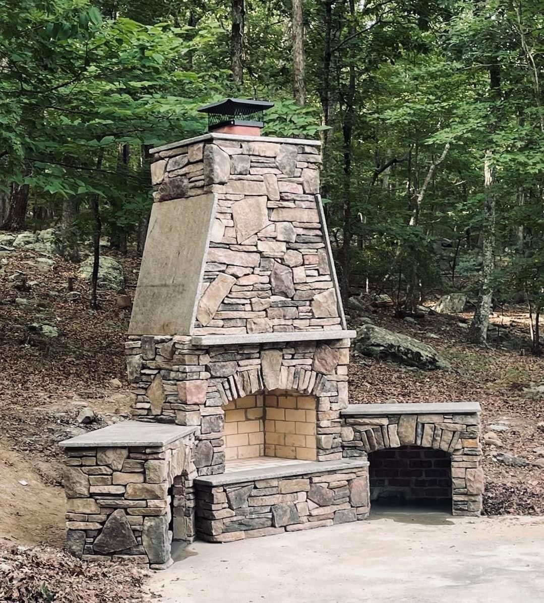 Stone outdoor fireplace with built-in grill and wood storage in a wooded area