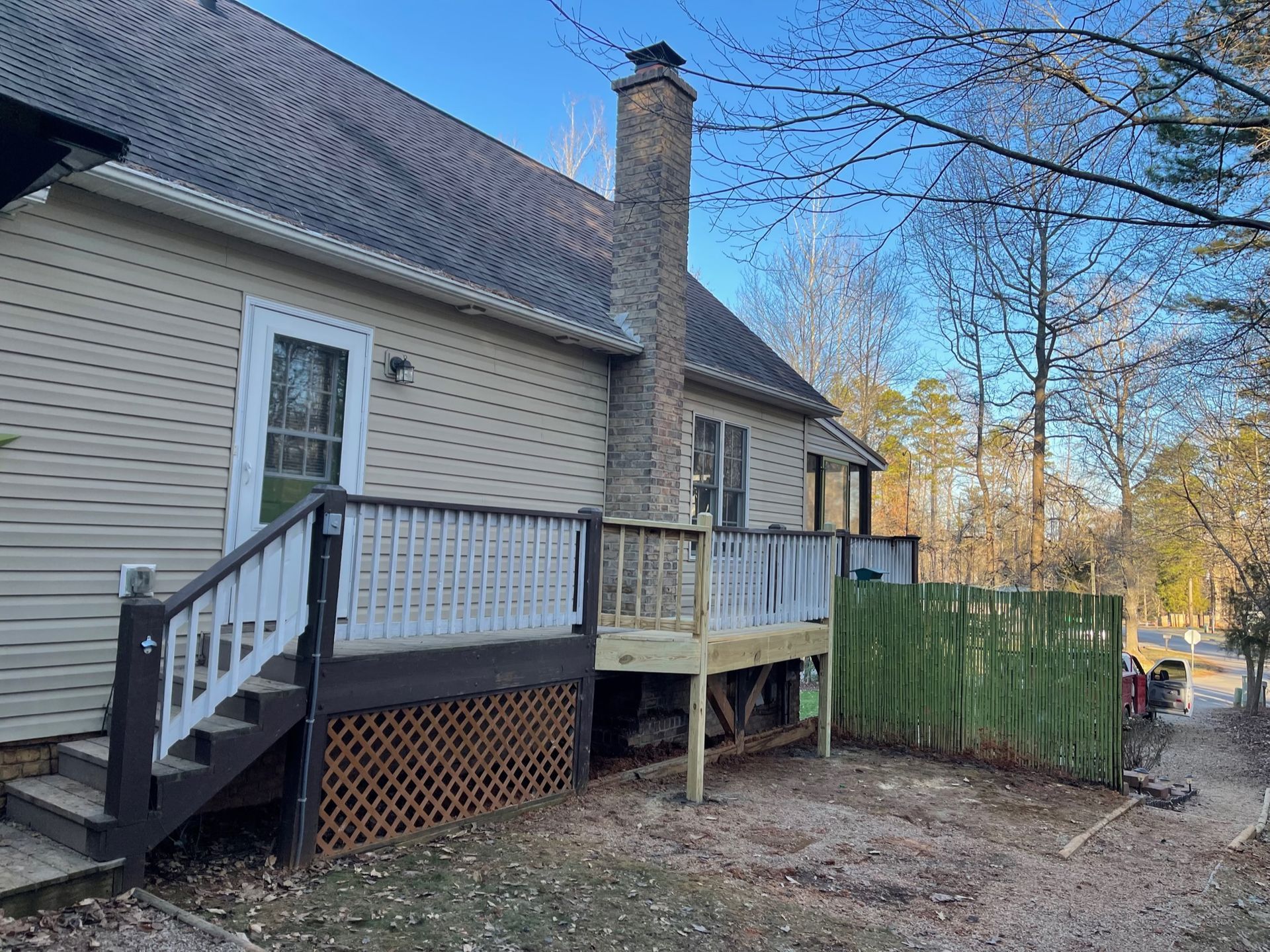 Back deck with white railing on beige house, brick chimney, and green fence in a wooded yard