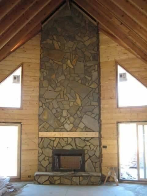 Cabin interior with a tall stone fireplace centered between two triangular windows and wooden walls.