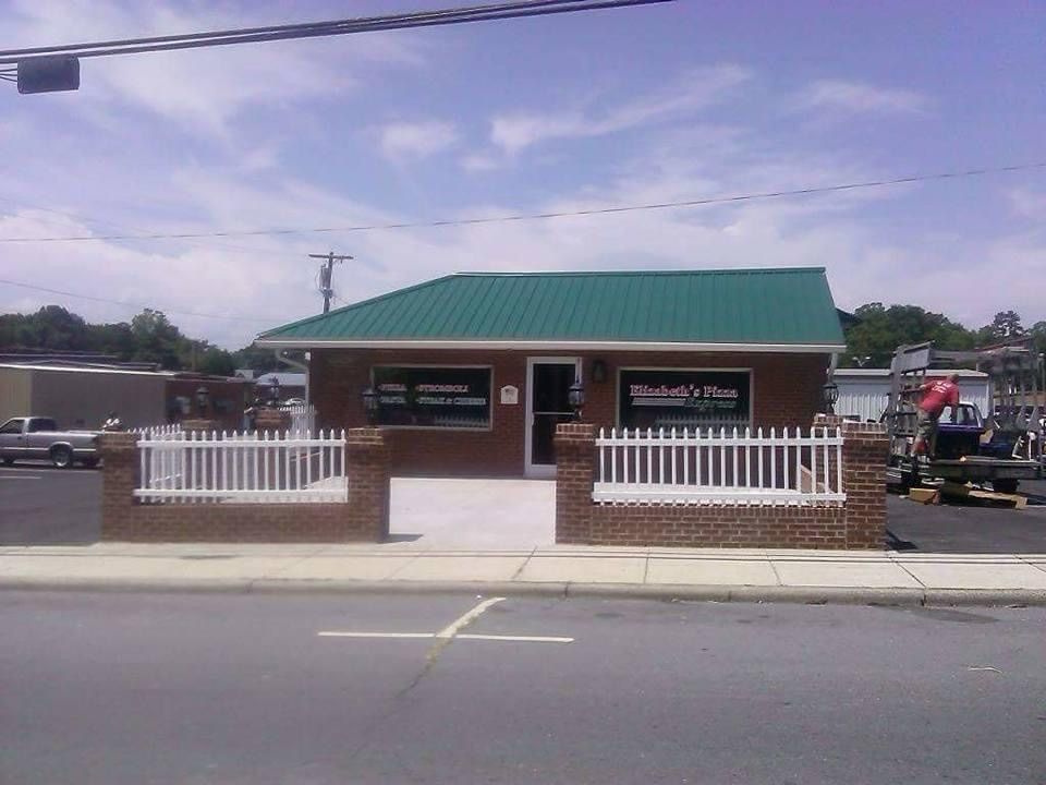 Small brick building with green roof and white fence beside a road under a cloudy sky
