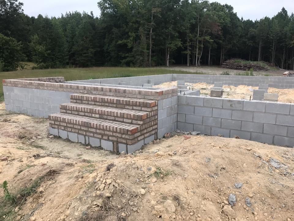 Partially built cinder block foundation with brick steps on a sandy lot, trees in the background