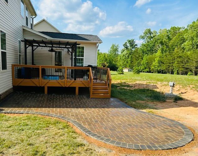 Backyard wooden deck with stairs beside a house and a curved brick patio leading to a grassy yard