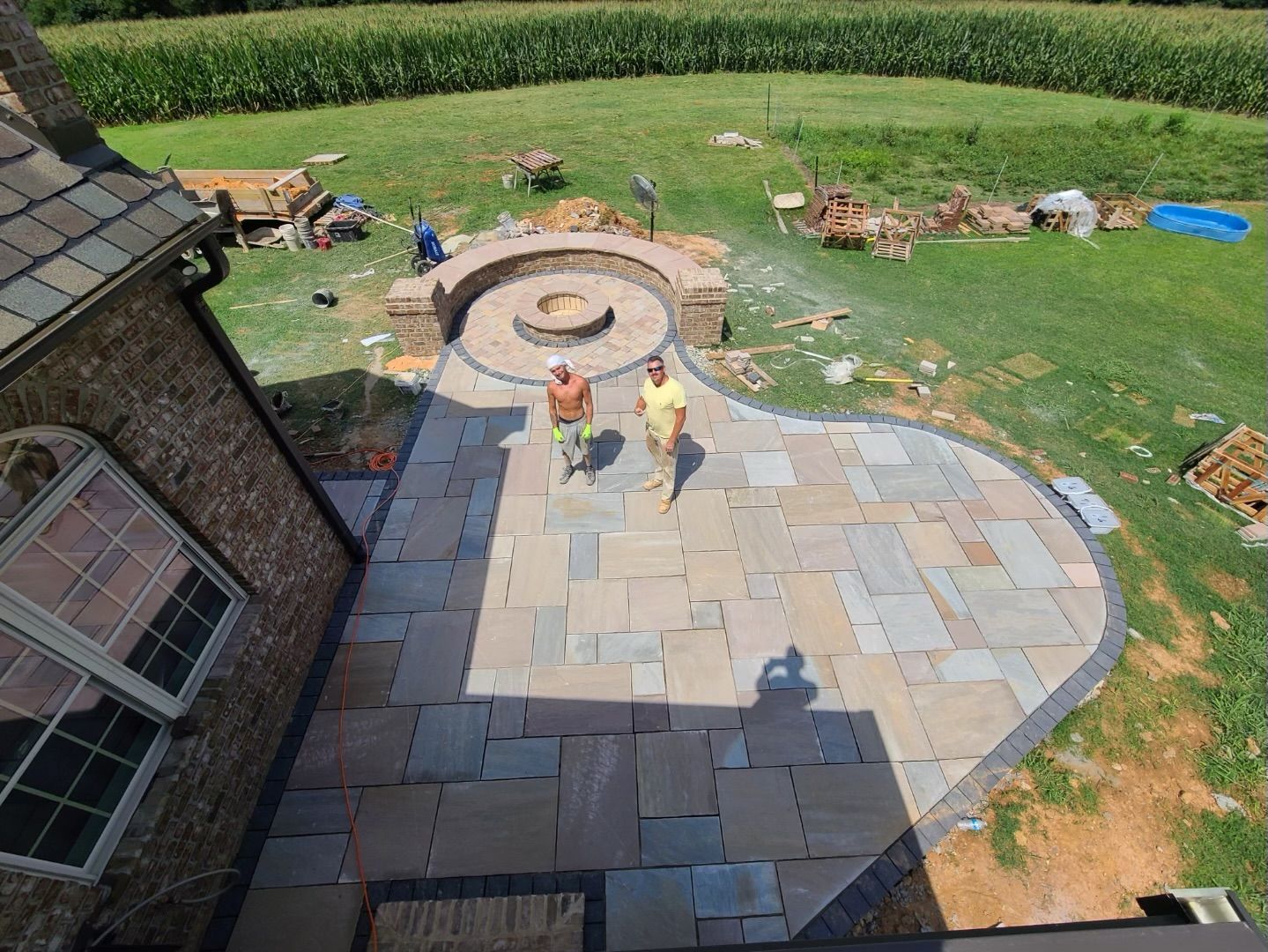 Backyard patio with workers installing stone paving beside a circular fire pit and lawn