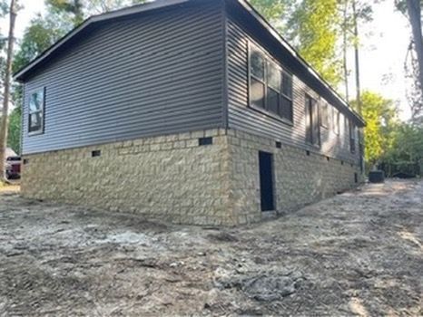 Side view of a gray house on a stone foundation in a wooded area with a dirt driveway