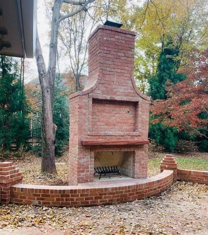 Brick outdoor fireplace with chimney, set in a curved brick wall amid autumn trees and fallen leaves
