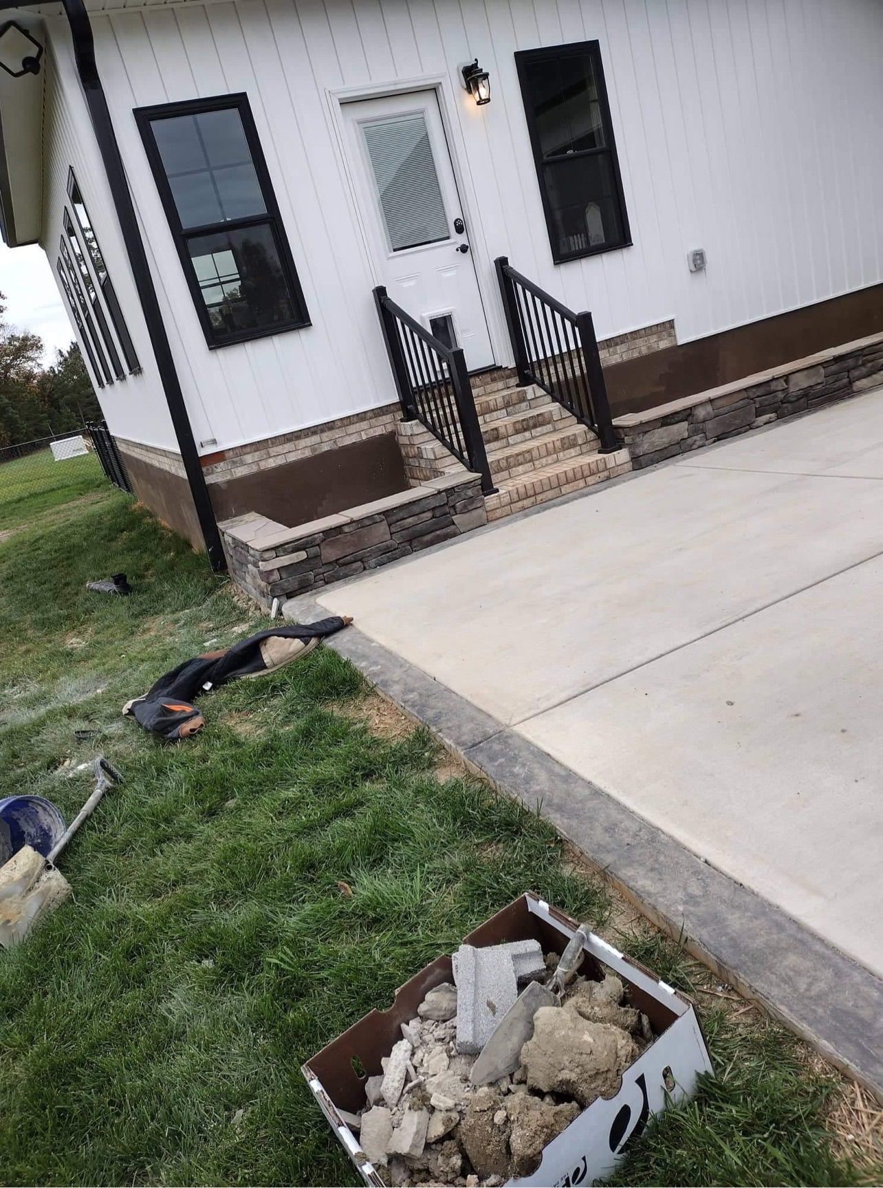 Tilted view of a white house entrance, driveway, grass, and construction debris in a box.