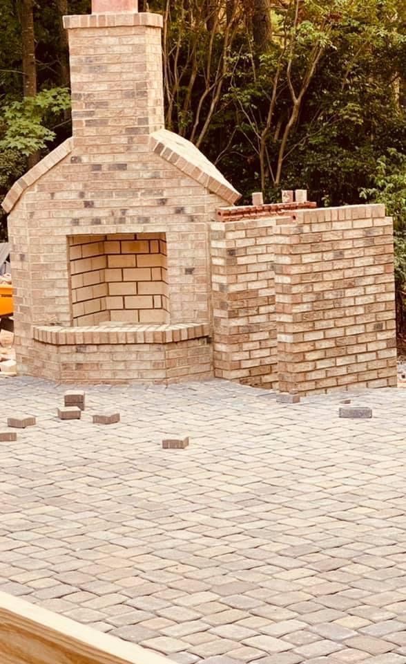 Brick outdoor fireplace with chimney on a paved patio, surrounded by trees.