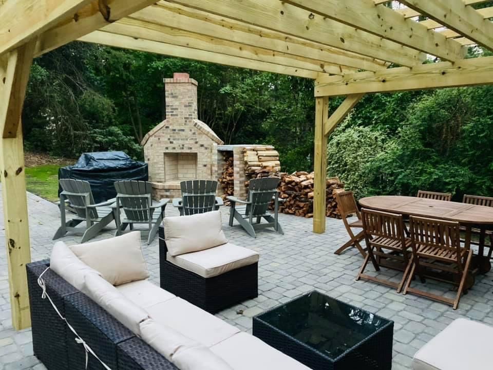 Covered patio with outdoor seating, fire pit, dining table, and stone outdoor fireplace in a wooded backyard
