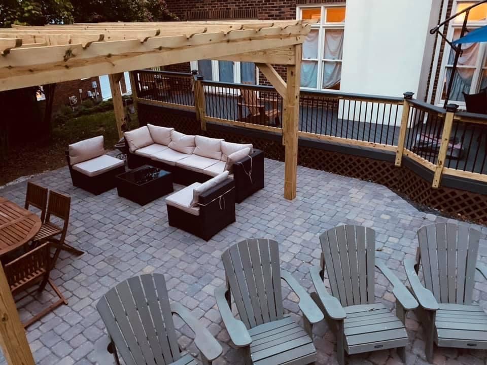 Backyard deck with wicker sectional seating, pergola, and several gray lounge chairs on a stone patio.