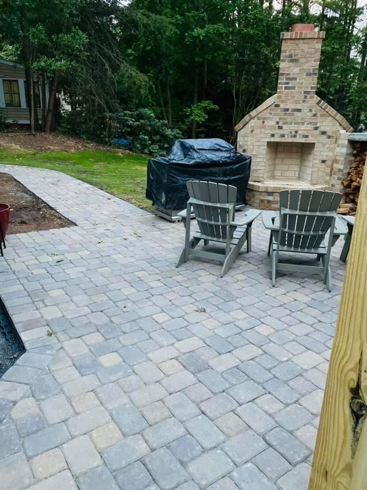 Patio with paver stones, two Adirondack chairs, a table, grill, and brick outdoor fireplace surrounded by trees