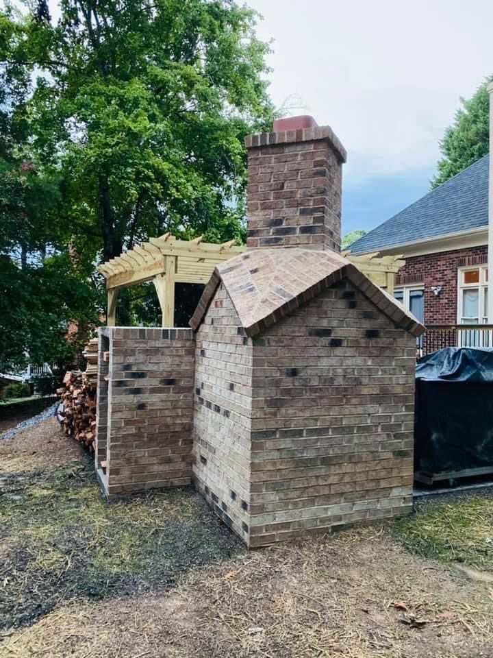 Brick outdoor fireplace and chimney beside a small shed in a backyard, with stacked firewood and trees.