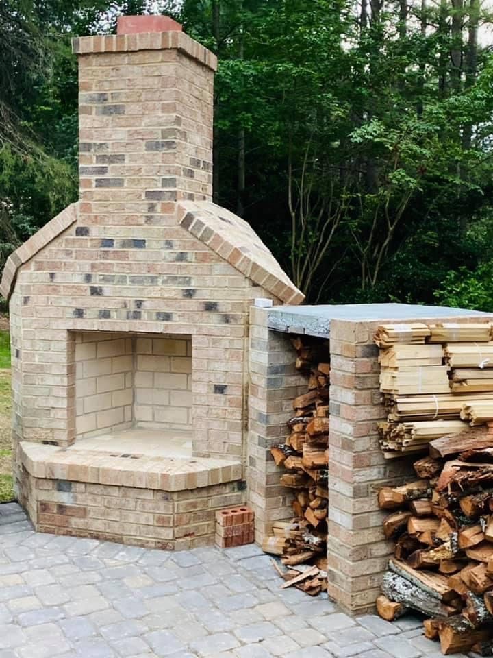 Brick outdoor fireplace beside a woodpile on a stone patio, with trees in the background