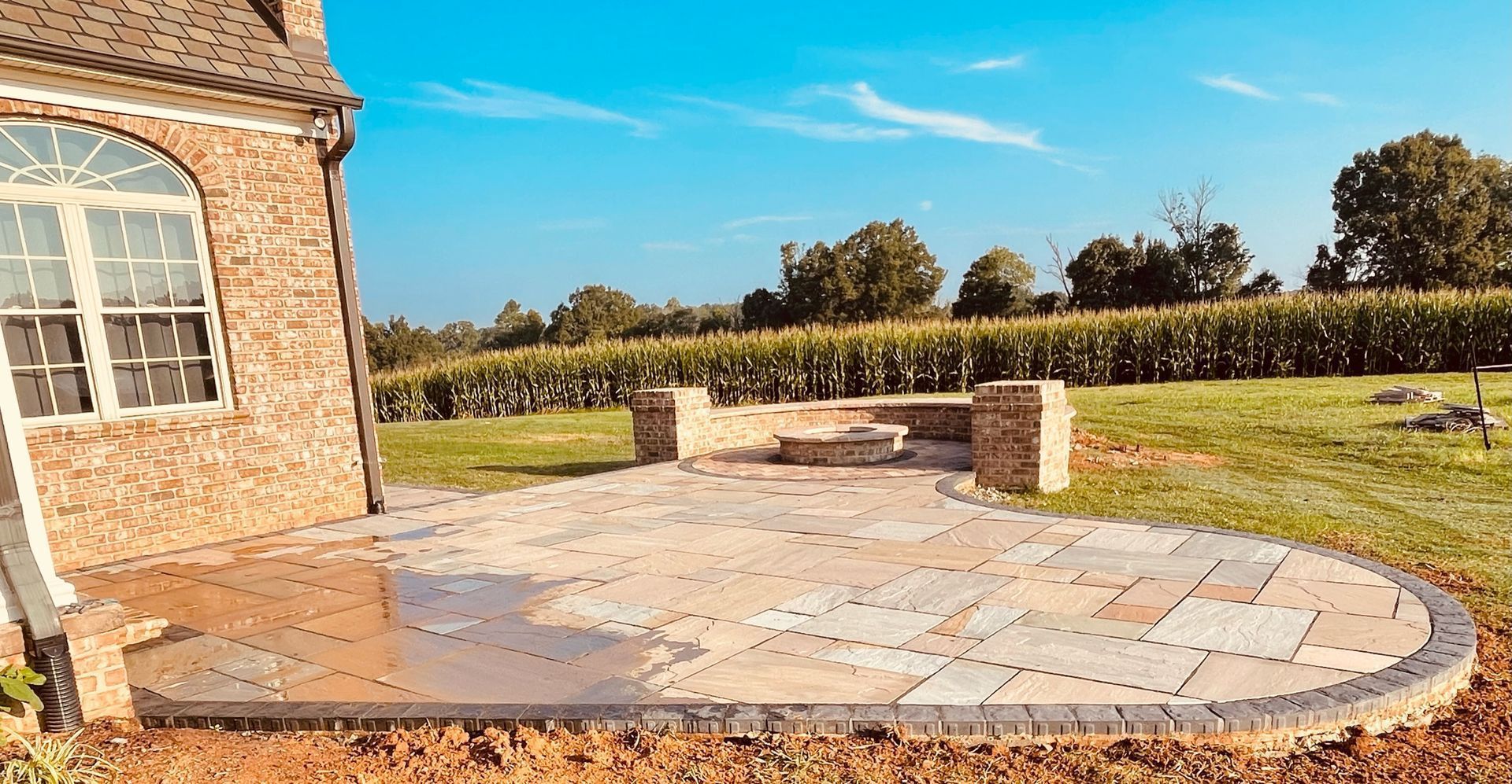 Stone patio beside a brick house, with a low stone wall and green fields under a clear blue sky.