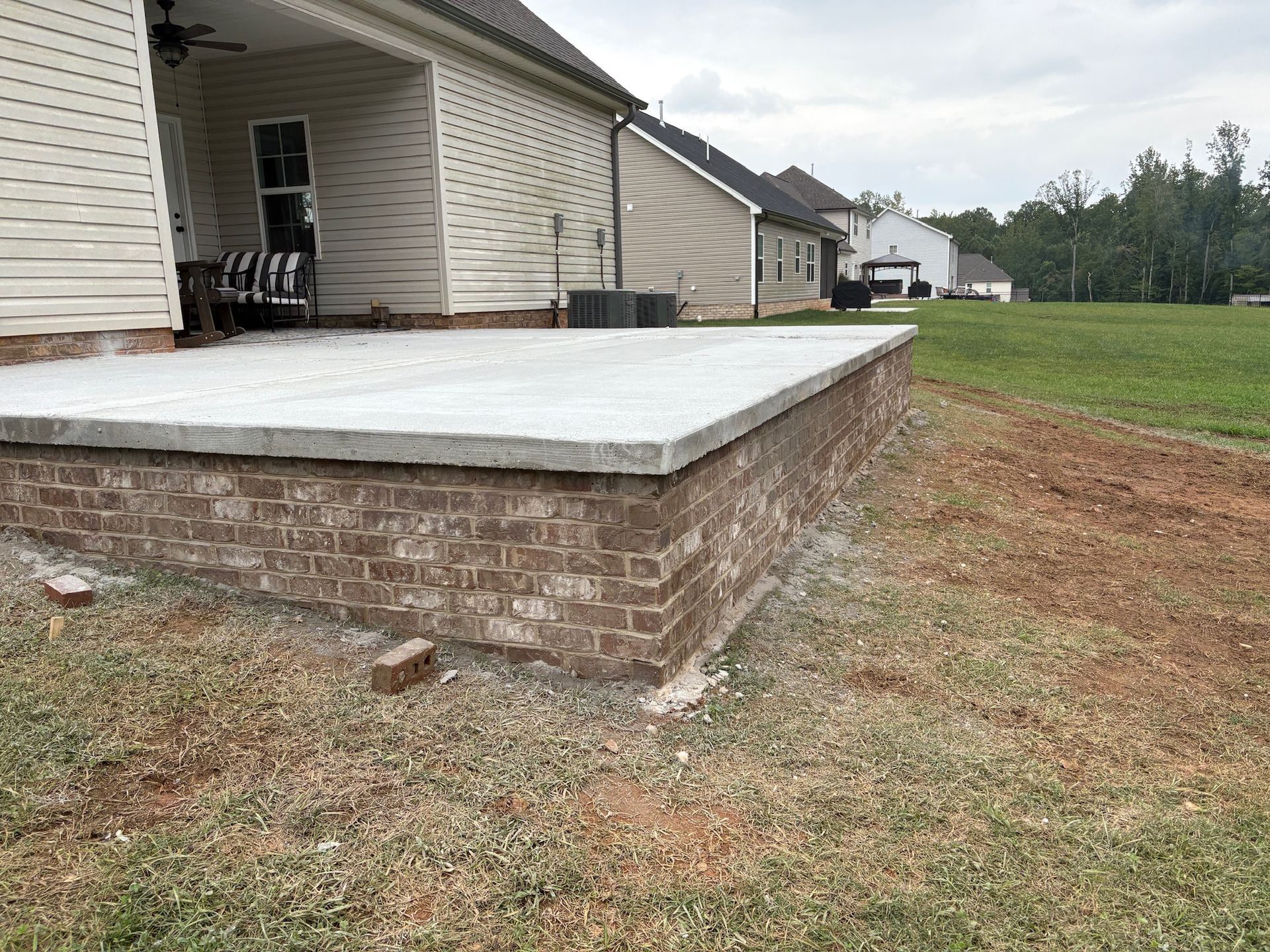 Raised concrete patio beside a beige house with a brick foundation and grassy yard.