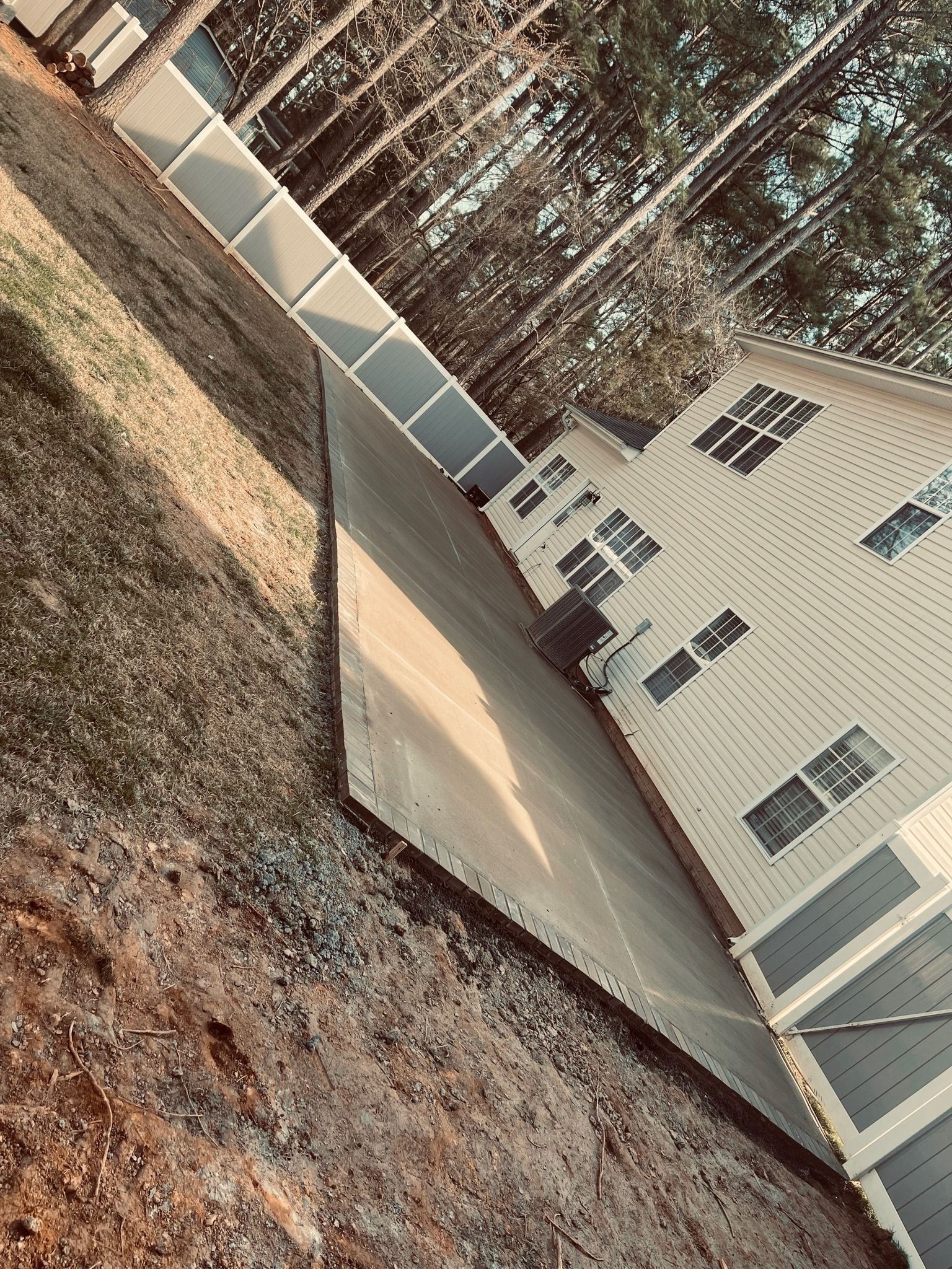 Tilted view of a beige house with white trim and a long concrete patio beside a wooded yard.