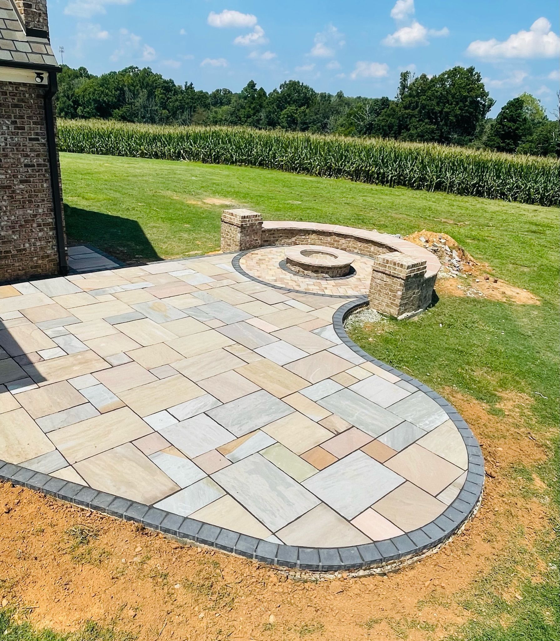 Curved stone patio with a built-in fire pit beside a lawn and hedge under a sunny sky