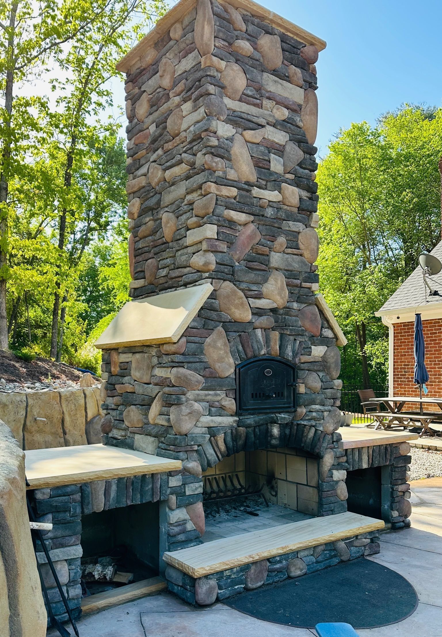 Stone outdoor fireplace with tall chimney and built-in benches in a wooded backyard
