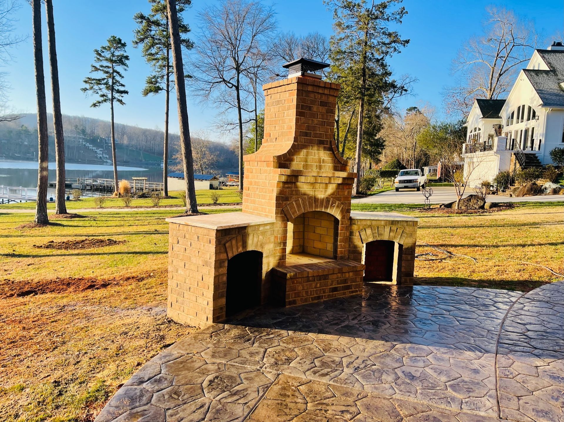Stone outdoor fireplace on a lakeside patio with trees and houses in the background