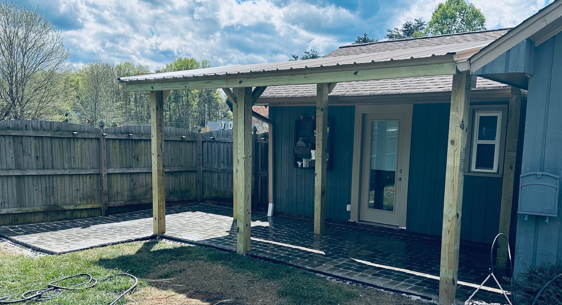Small backyard porch with weathered wooden posts, a blue house wall, and a fenced yard under a cloudy sky