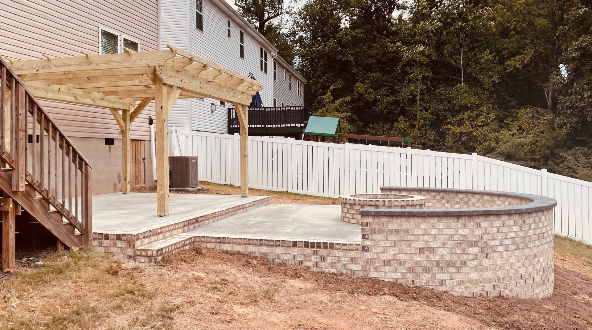 Backyard patio with wooden pergola, stairs, and raised stone fire pit beside a house and white fence