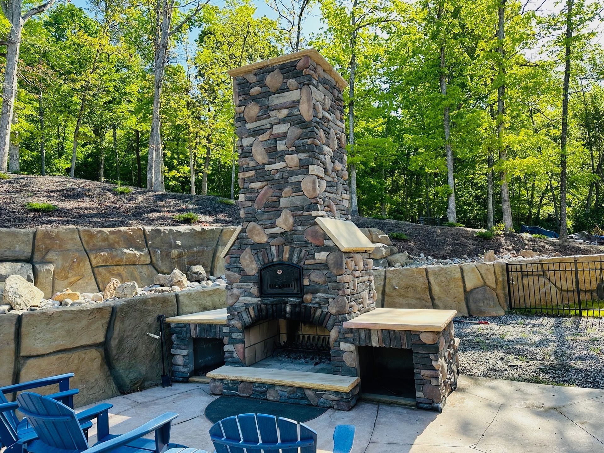 Stone outdoor fireplace with built-in benches and blue chairs in a wooded yard