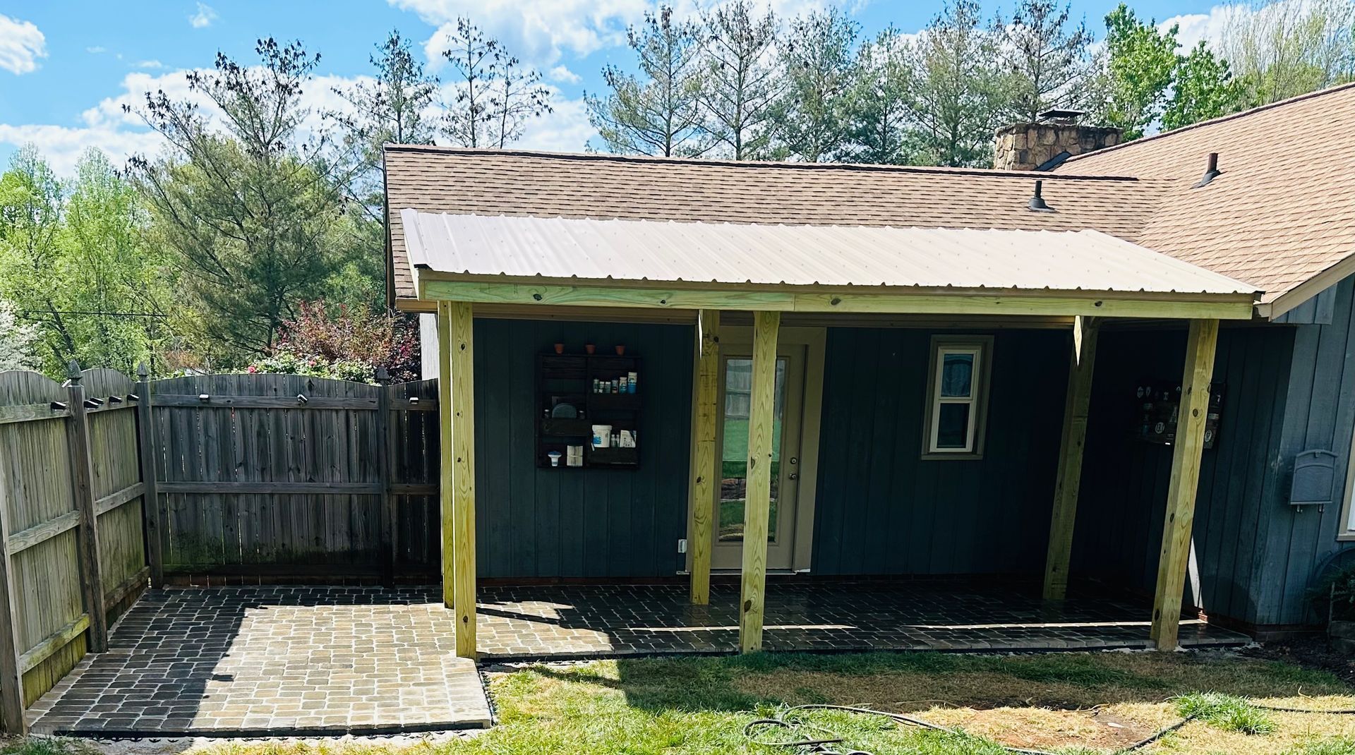 Small wooden porch with green posts on a blue house, beside a fenced backyard with trees.