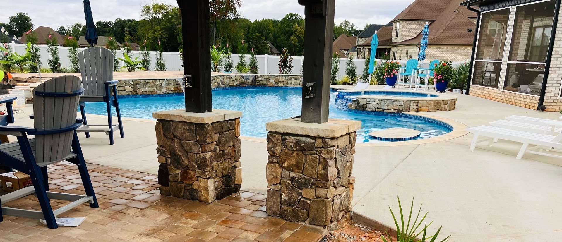 Covered poolside patio with stone columns, blue chairs, and a hot tub beside a fenced swimming pool.