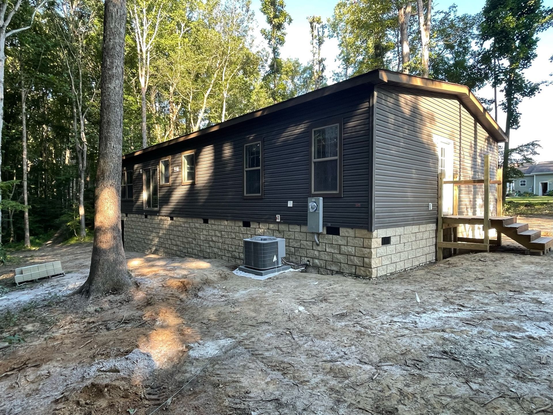 Small dark cabin with tan brick base and wooden steps in a wooded area, with a window AC unit.