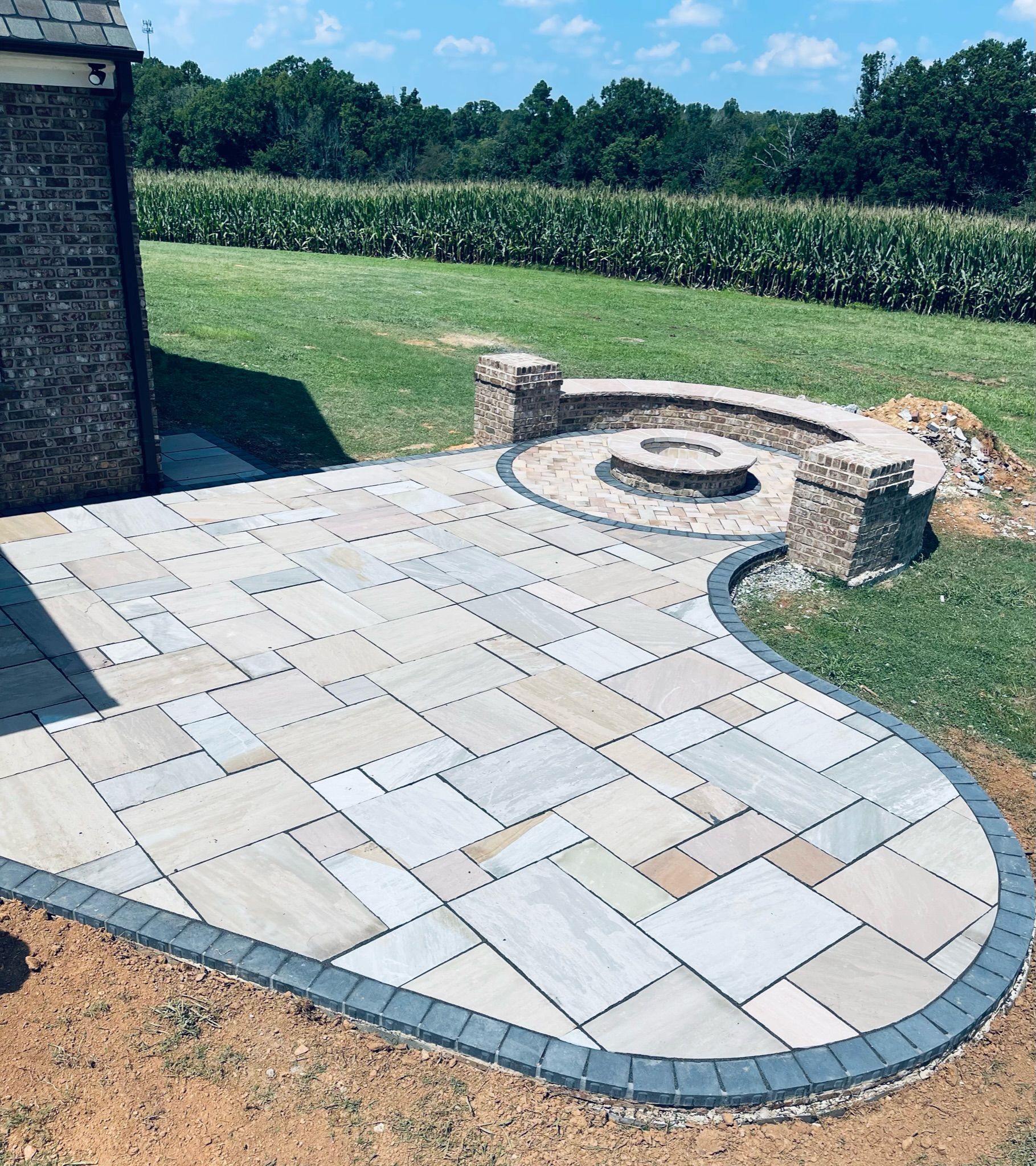 Stone patio with curved edging and a circular fire pit beside a grassy lawn and hedge.