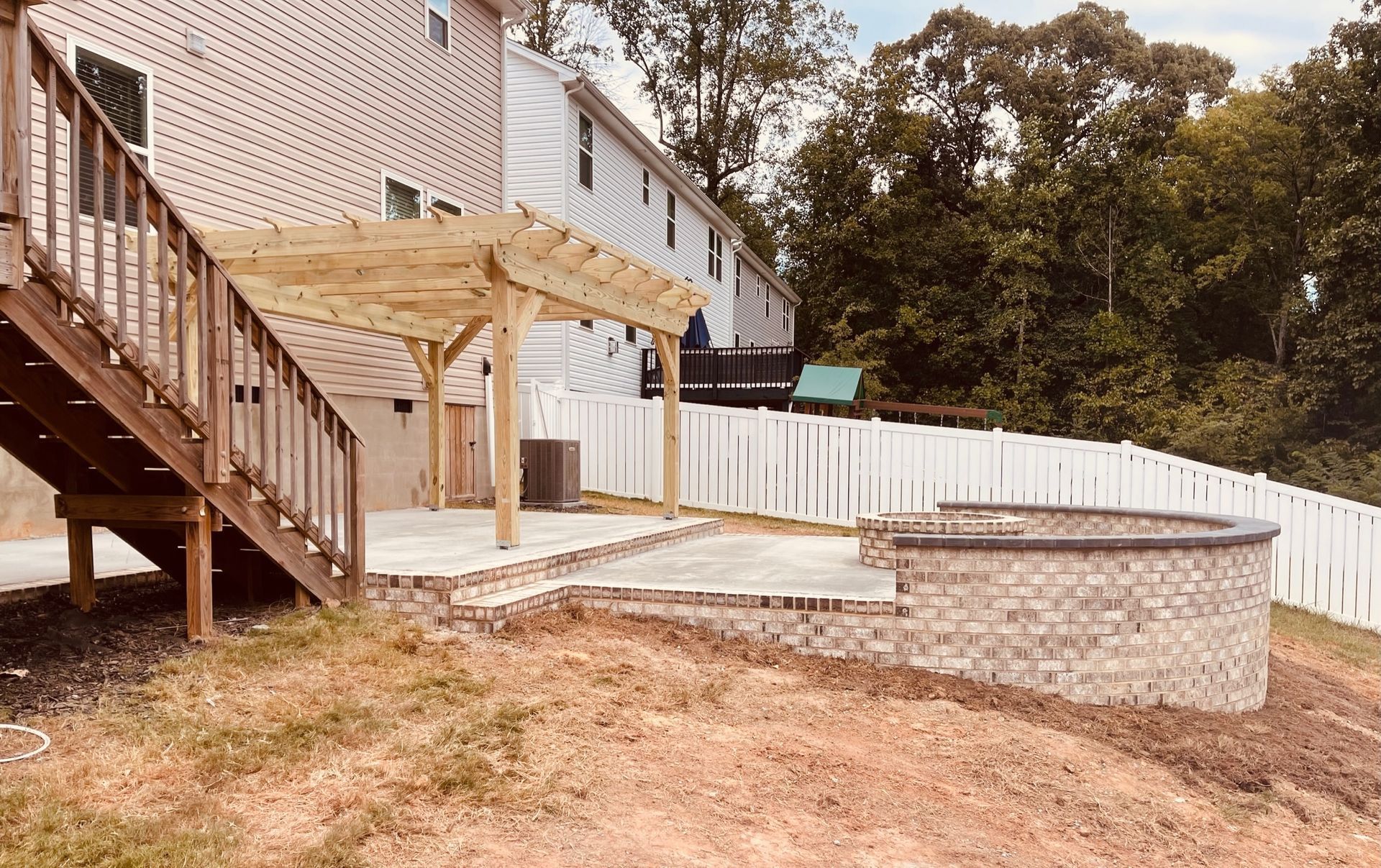 Backyard patio with stone retaining wall, wooden deck stairs, and white fence beside a house