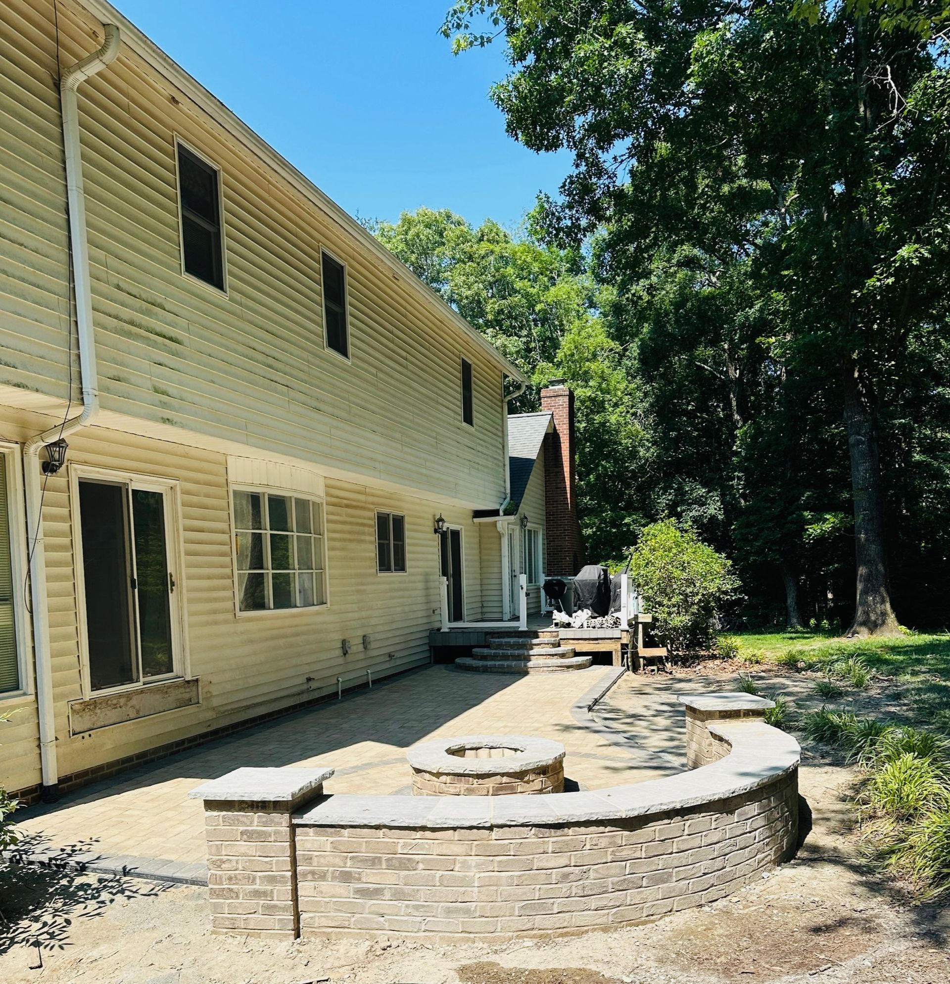 Backyard patio with curved stone seating wall beside a yellow house and trees.