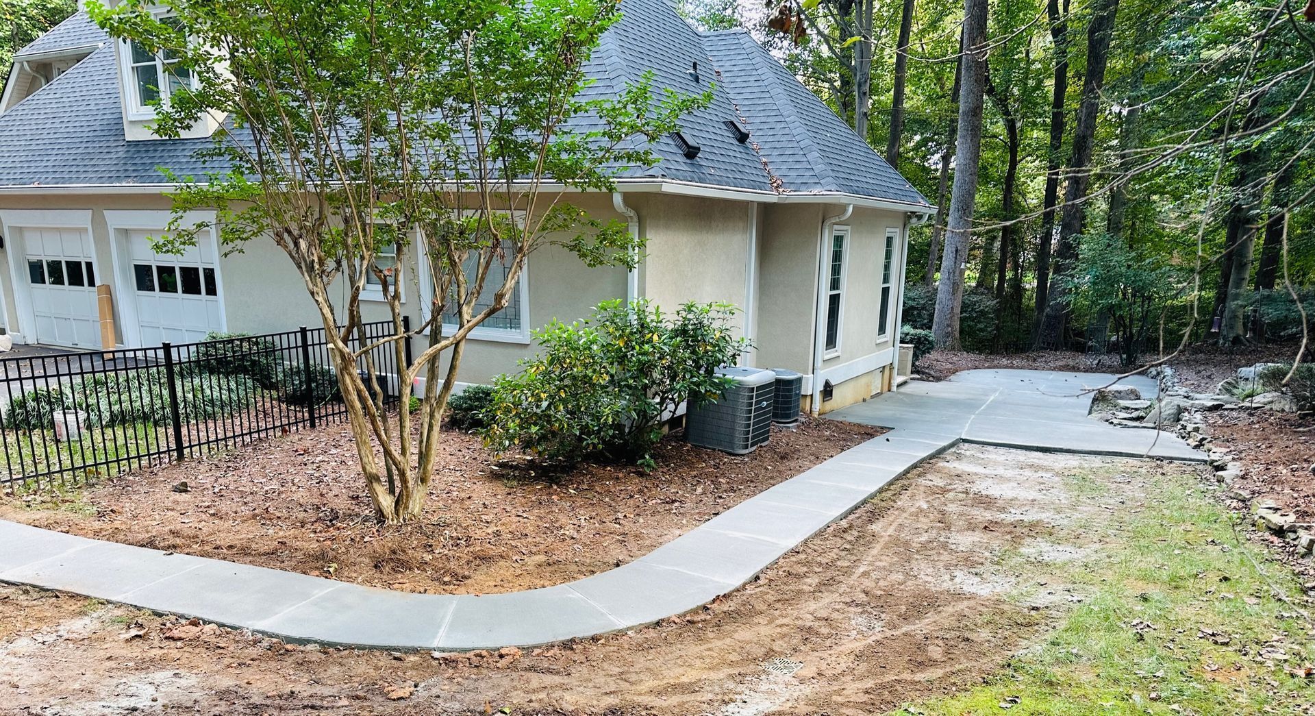 Small house with gray roof, white siding, and curving concrete walkway in a wooded yard.