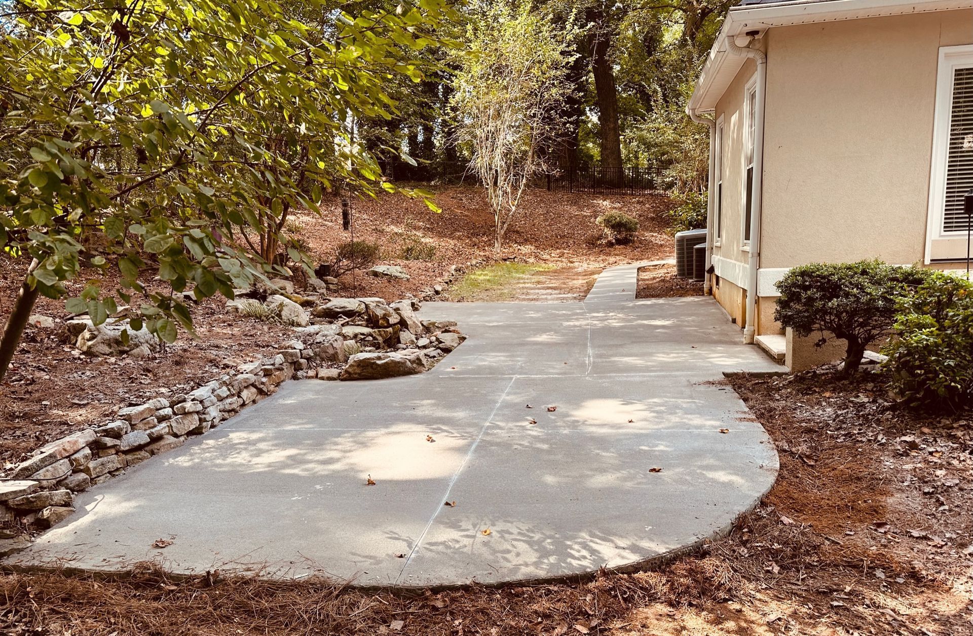 Curved concrete walkway beside a beige house, bordered by rocks and trees in a wooded yard.