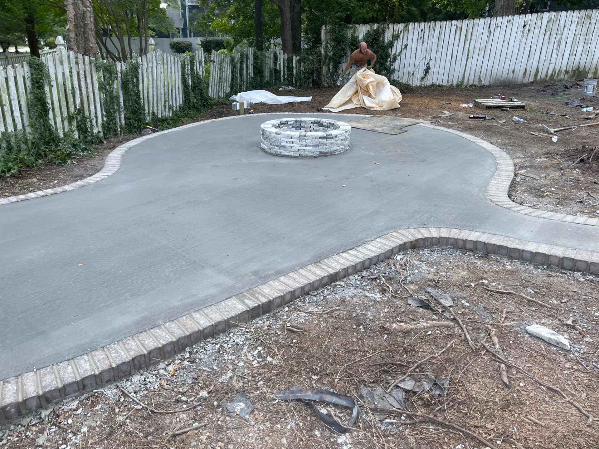 Concrete patio with a circular fire pit, bordered by a curved brick edge in a backyard.