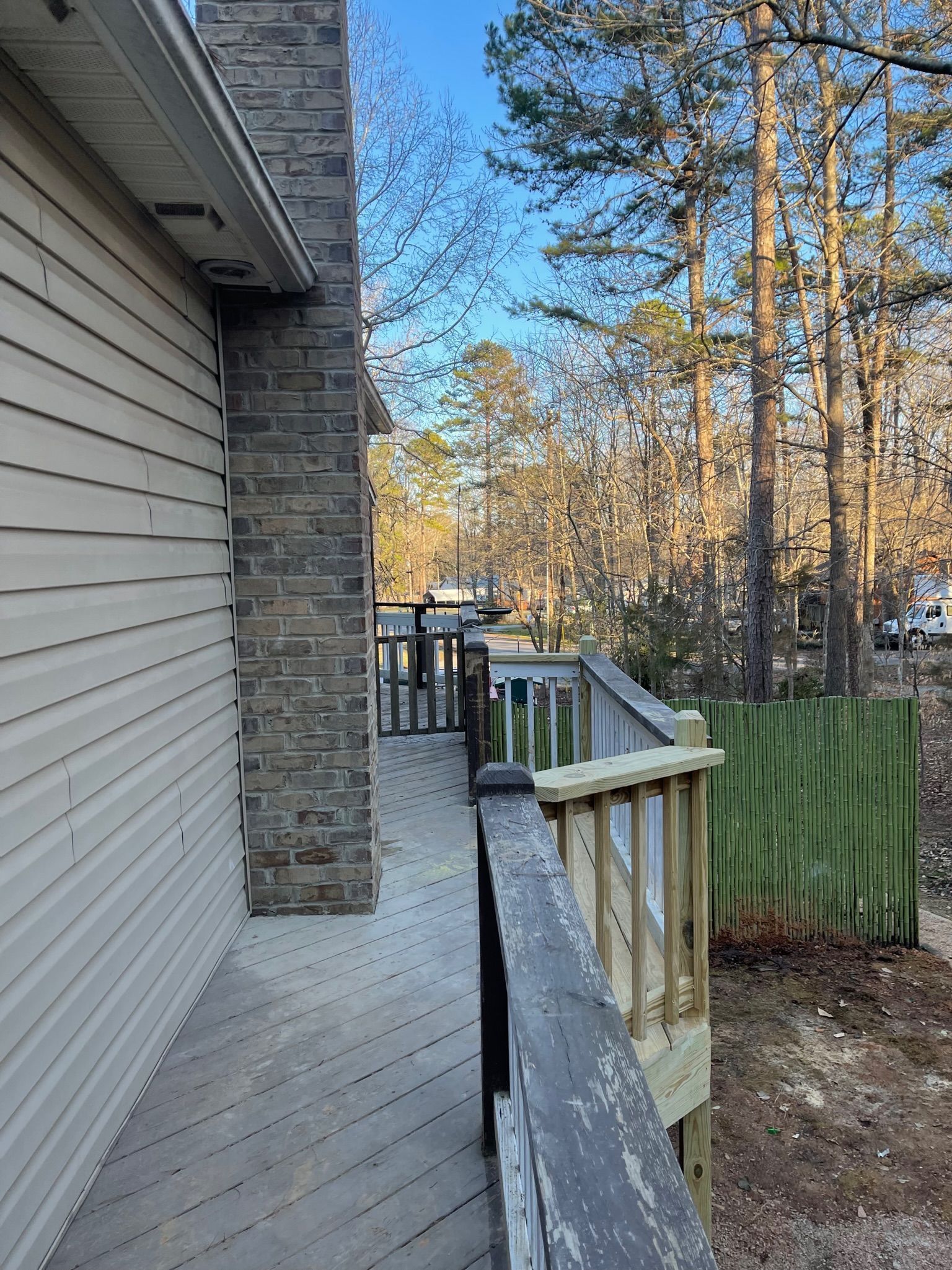 Side view of a house’s wooden deck beside a stone wall, with a railing and wooded yard beyond.