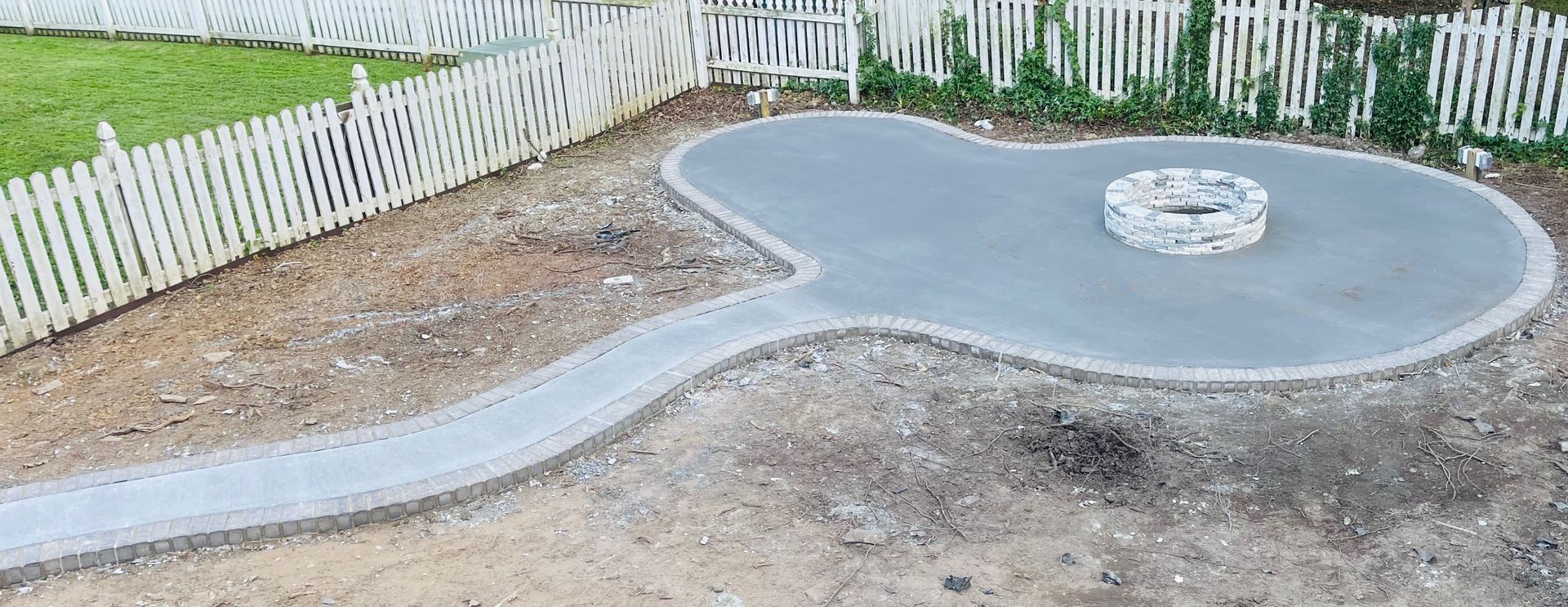 New concrete patio with curved walkway and round fire pit near a white picket fence