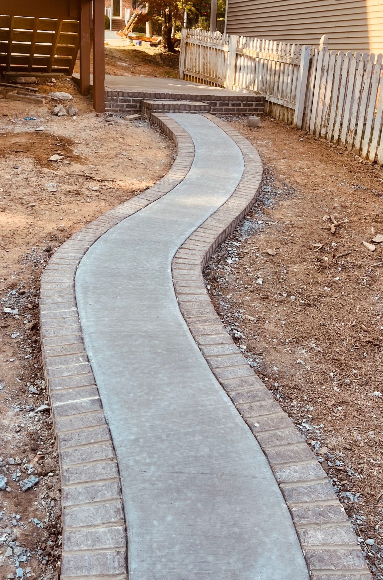 Curved concrete garden path with stone edging beside a wooden fence and dry mulch