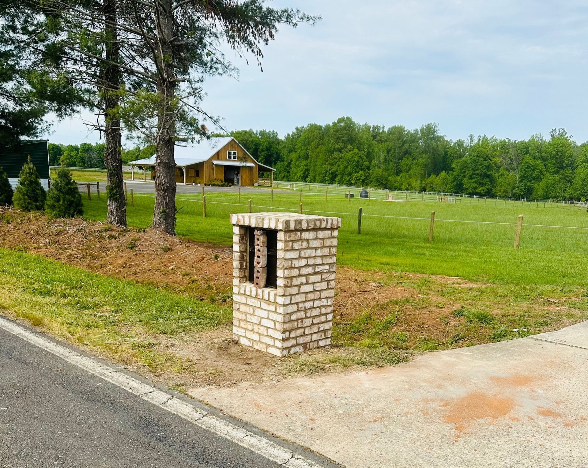 Brick mailbox by a rural road near a grassy field and house under trees.