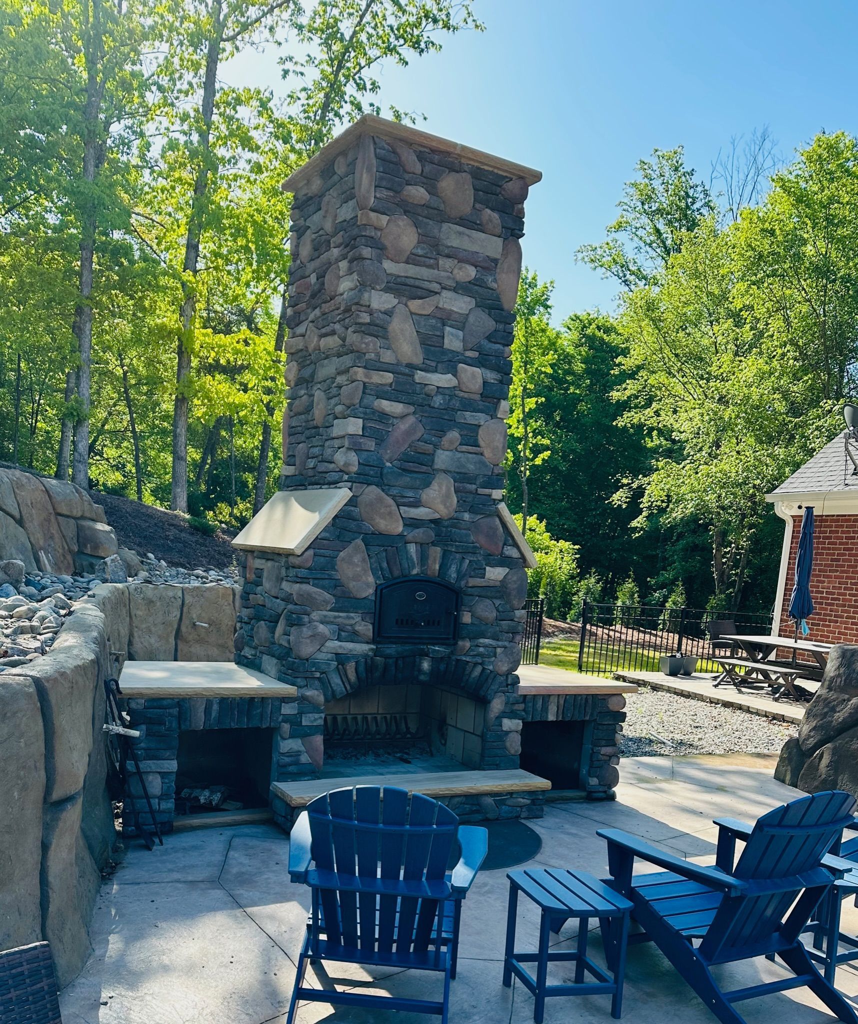 Stone outdoor fireplace with blue Adirondack chairs on a sunny patio, surrounded by trees.