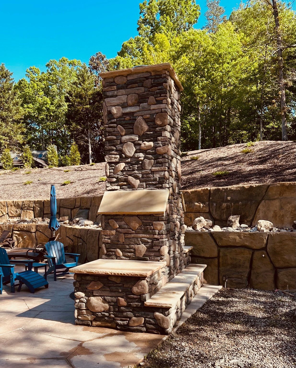 Stone outdoor fireplace with blue patio chairs in a wooded yard under bright sunlight